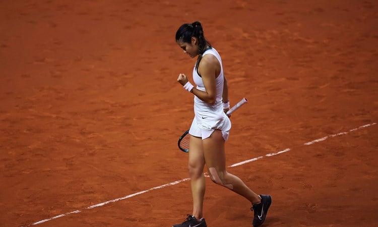 Emma Raducanu clenching her fist while stood on a clay court in a white tennis dress 