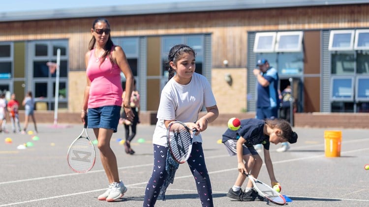 an image of a young girl holding a tennis racket and hitting a ball on court with other children and an older woman stood around her