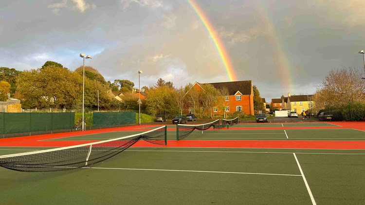 A picture of three tennis courts with houses behind them and a rainbow in the sky
