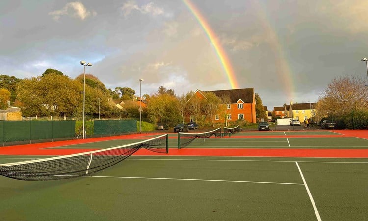 A picture of three tennis courts with houses behind them and a rainbow in the sky