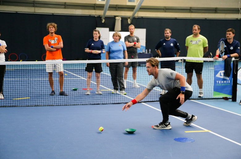 Prospective tennis coaches standing on a tennis court watching a tutor during an LTA on-court workshop