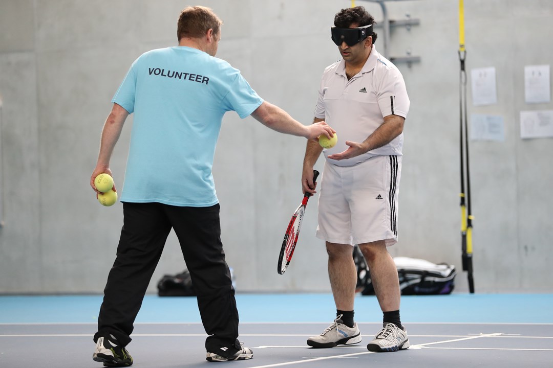 Volunteer hands a tennis ball to a visually impaired player