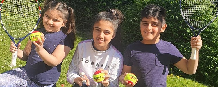 three kids smiling all holding a yellow and red tennis ball and two with tennis rackets in the air