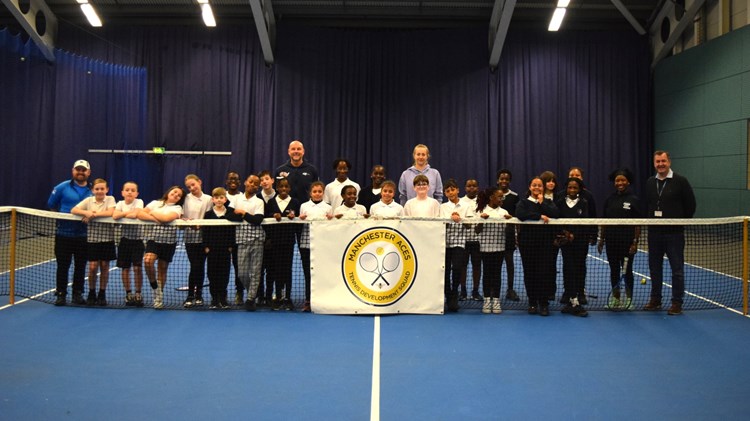 A group of children stand behind a tennis net on a blue tennis court. Behind the group in the middle stand two adults, a man left of centre and a woman right of centre, whilst at the far left a man stands in a blue tacksuit and on the far rihgt a man stands weating a black jumper and trousers