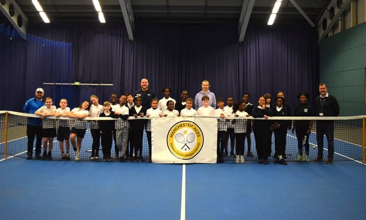 A group of children stand behind a tennis net on a blue tennis court. Behind the group in the middle stand two adults, a man left of centre and a woman right of centre, whilst at the far left a man stands in a blue tacksuit and on the far rihgt a man stands weating a black jumper and trousers