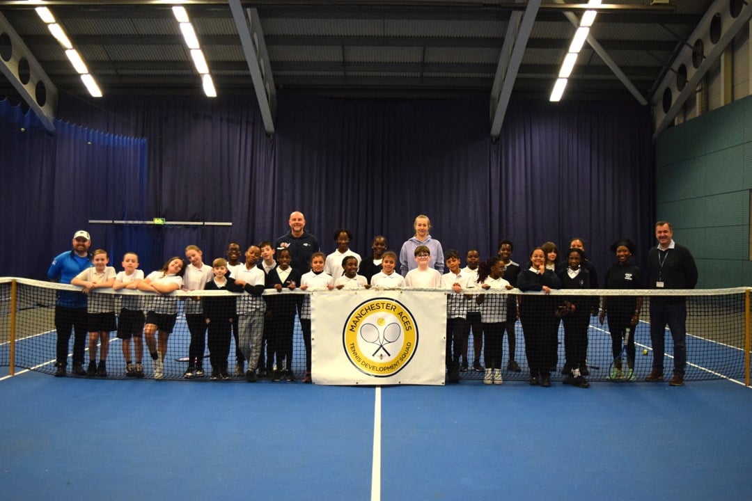 A group of children stand behind a tennis net on a blue tennis court. Behind the group in the middle stand two adults, a man left of centre and a woman right of centre, whilst at the far left a man stands in a blue tacksuit and on the far rihgt a man stands weating a black jumper and trousers