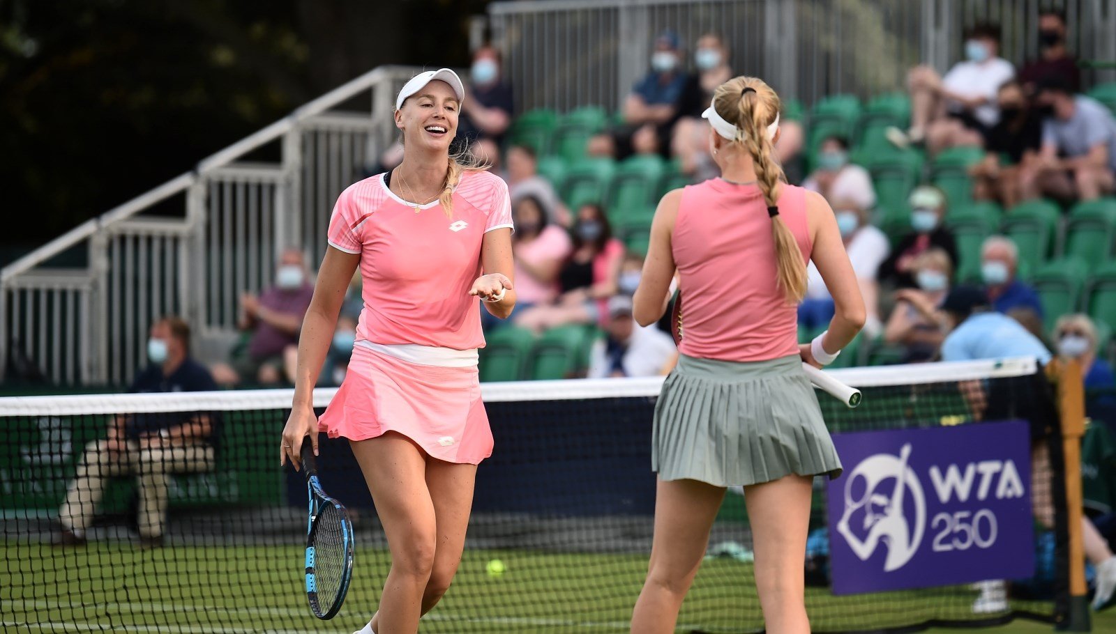Two women wearing pink shirts on a grass tennis court. One woman is facing the camera running towards the other woman with a smile on her face