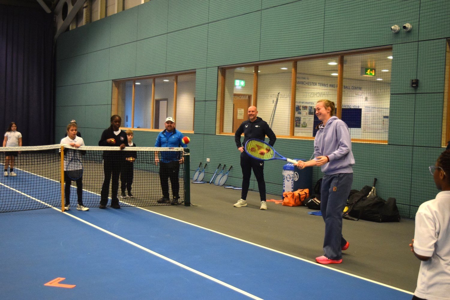 A woman wearing a light purple hoodi and tracksuit bottoms smiles whilst holding a tennis racket. She is stood on the edge of a tennis court, and in the background, two men and a number of schoolchildren watch the woman as she hits a ball.