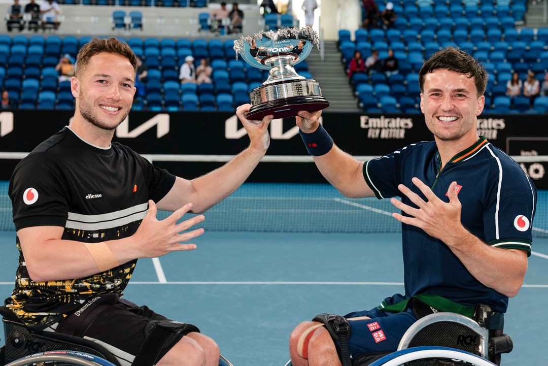 Alfie Hewett and Gordon Reid holding their Australian Open trophy while showing five fingers on their hands to signify five successive Aus Open titles