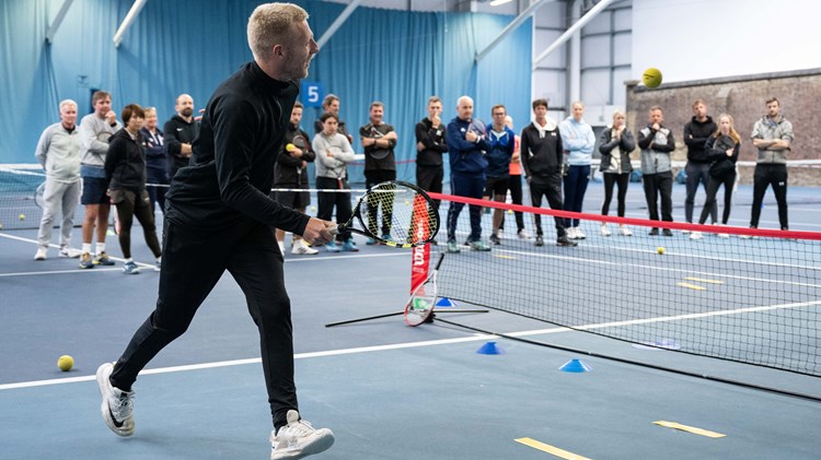 Male coach on mini tennis court demonstrating drill to crowd