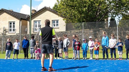 Children lined up on an outdoor tennis court with a coach standing in front of them.