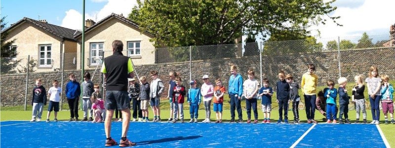 Children lined up on an outdoor tennis court with a coach standing in front of them.