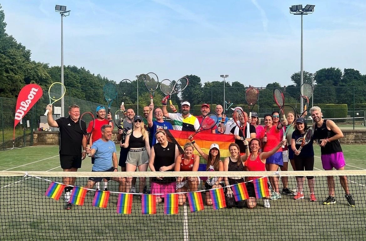 A large group of people standing on a tennis court, posing with tennis rackets dressed in brightly coloured clothing, behind a net draped with rainbow flags