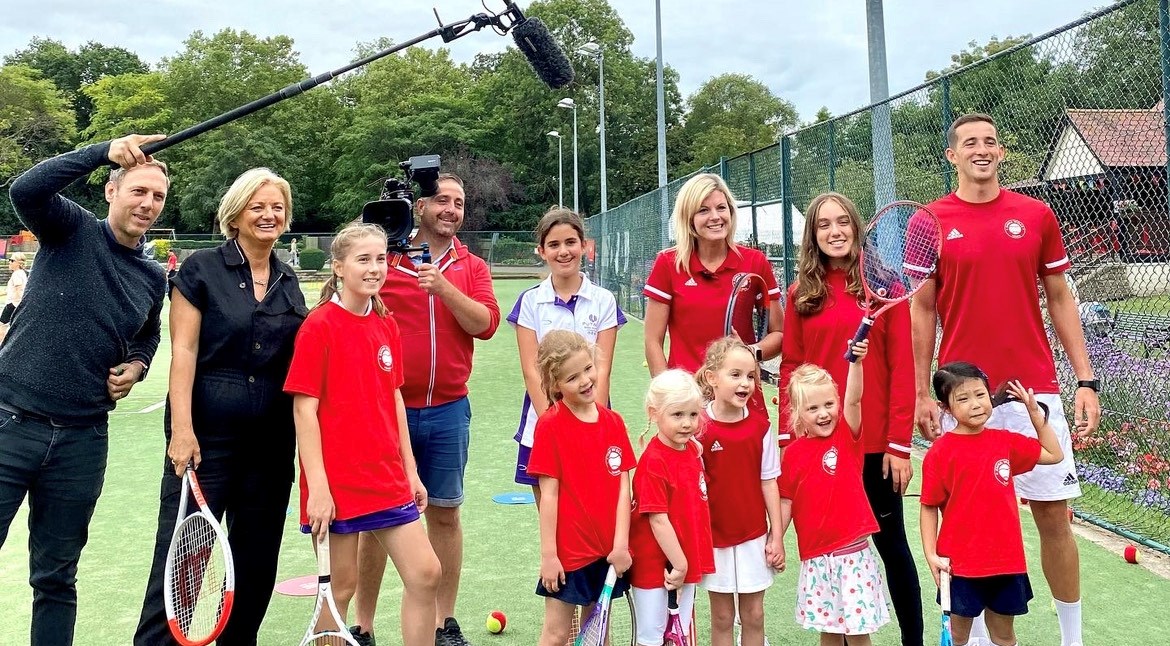 A group of children and adults posing with tennis rackets, with a man holding a large microphone and a man holding a tv camera stood next to them