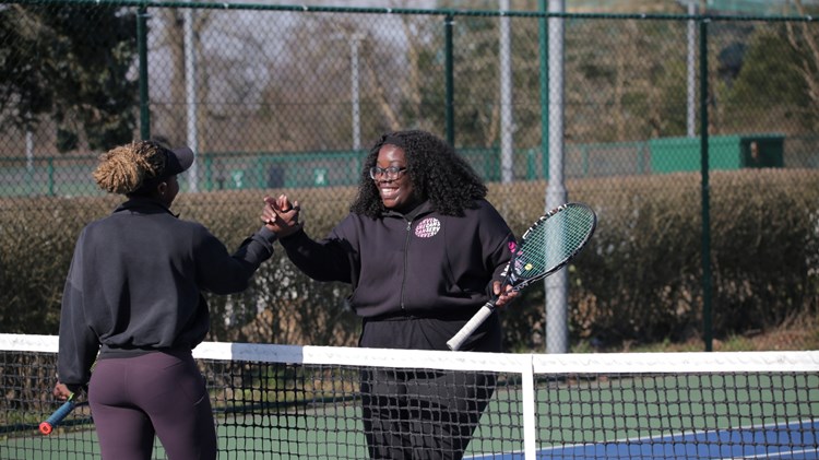Two women shake hands over the top of a tennis net, each holding a tennis racket in their left hand. Tennis court markings and fecing are visible in the background.