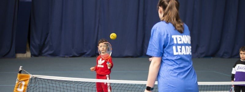 A girl in a tennis leader t-shirt throwing a ball to a young boy holding a tennis racket. 