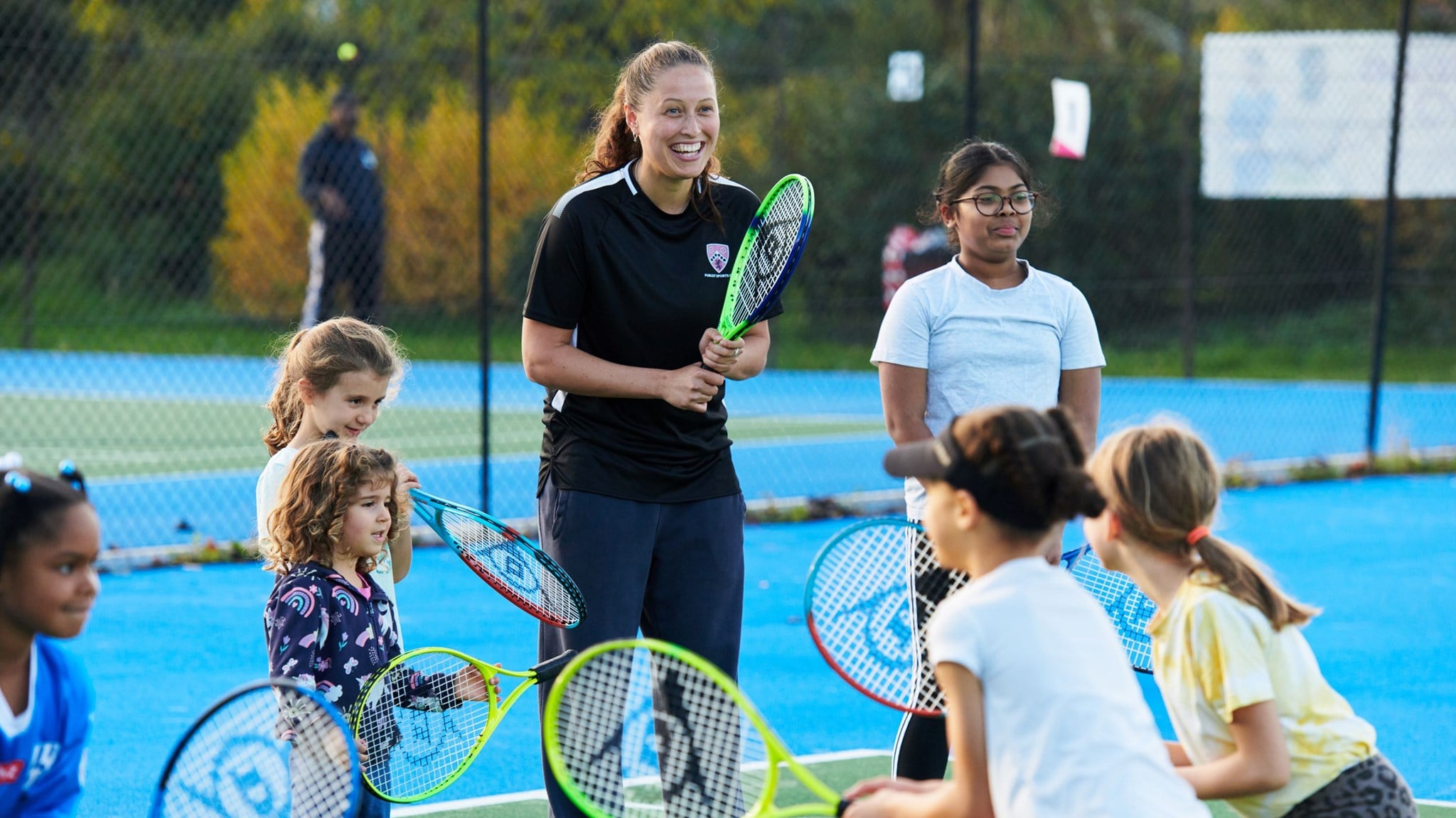 A coach on court, holding a racket and smiling while a group of young players complete a drill with rackets in front of her