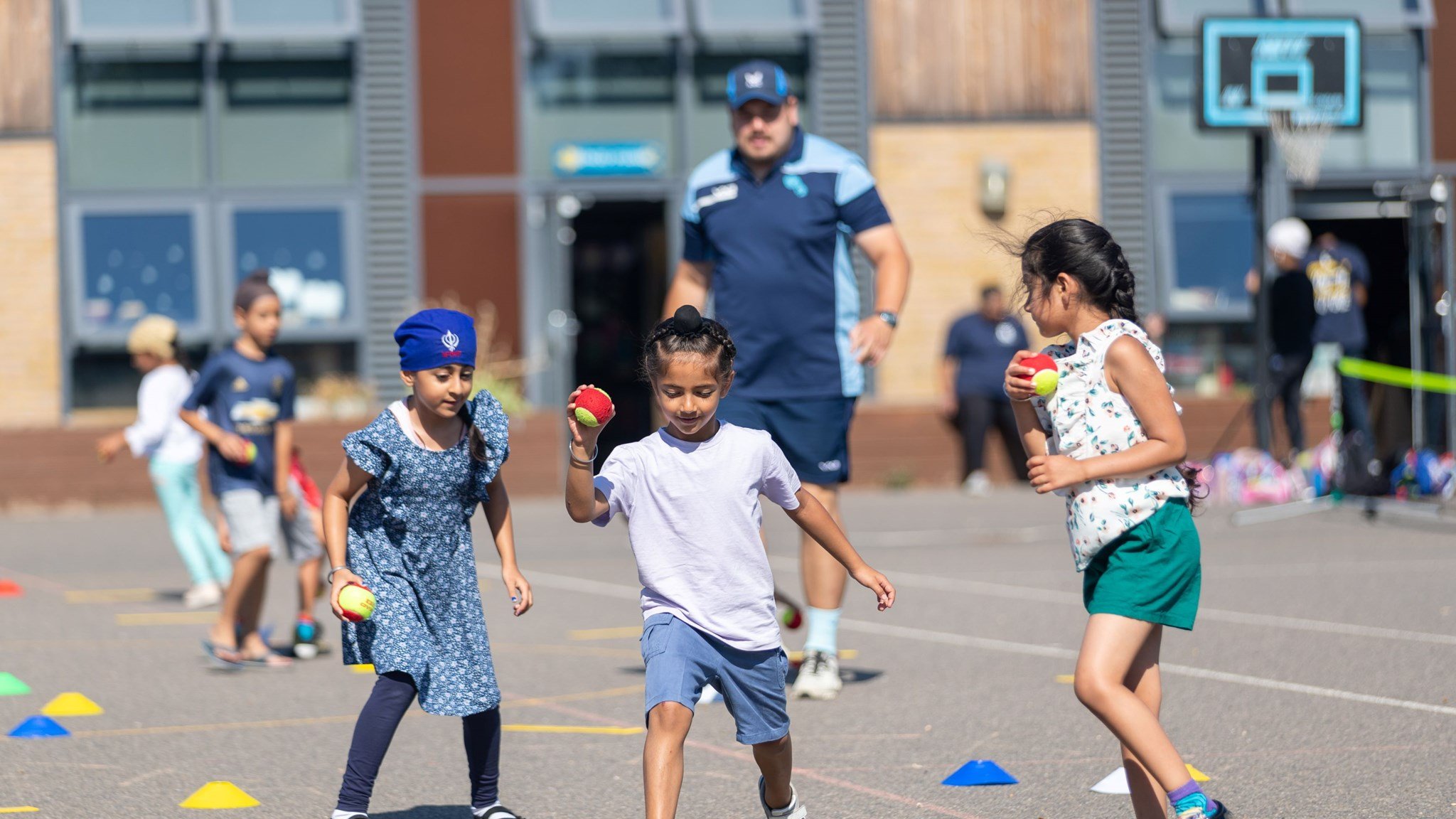 Three young kids playing with tennis balls and cones on the ground while their coach watches on