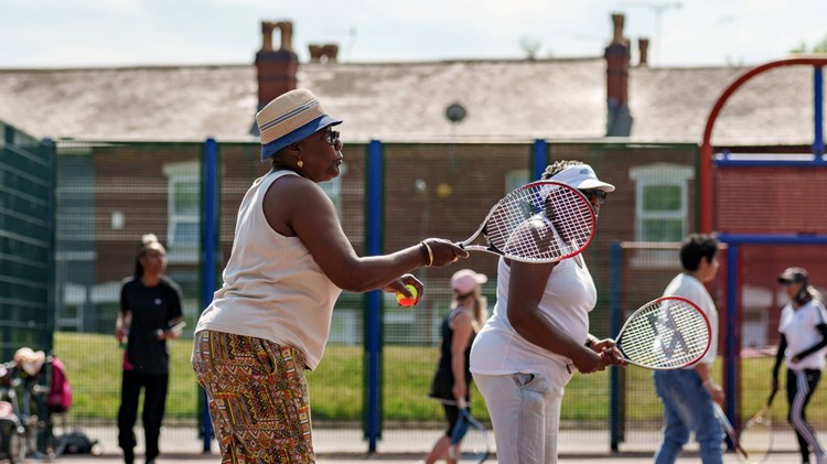 An image of a group of ladies holding tennis rackets and balls on a concrete tennis court group session