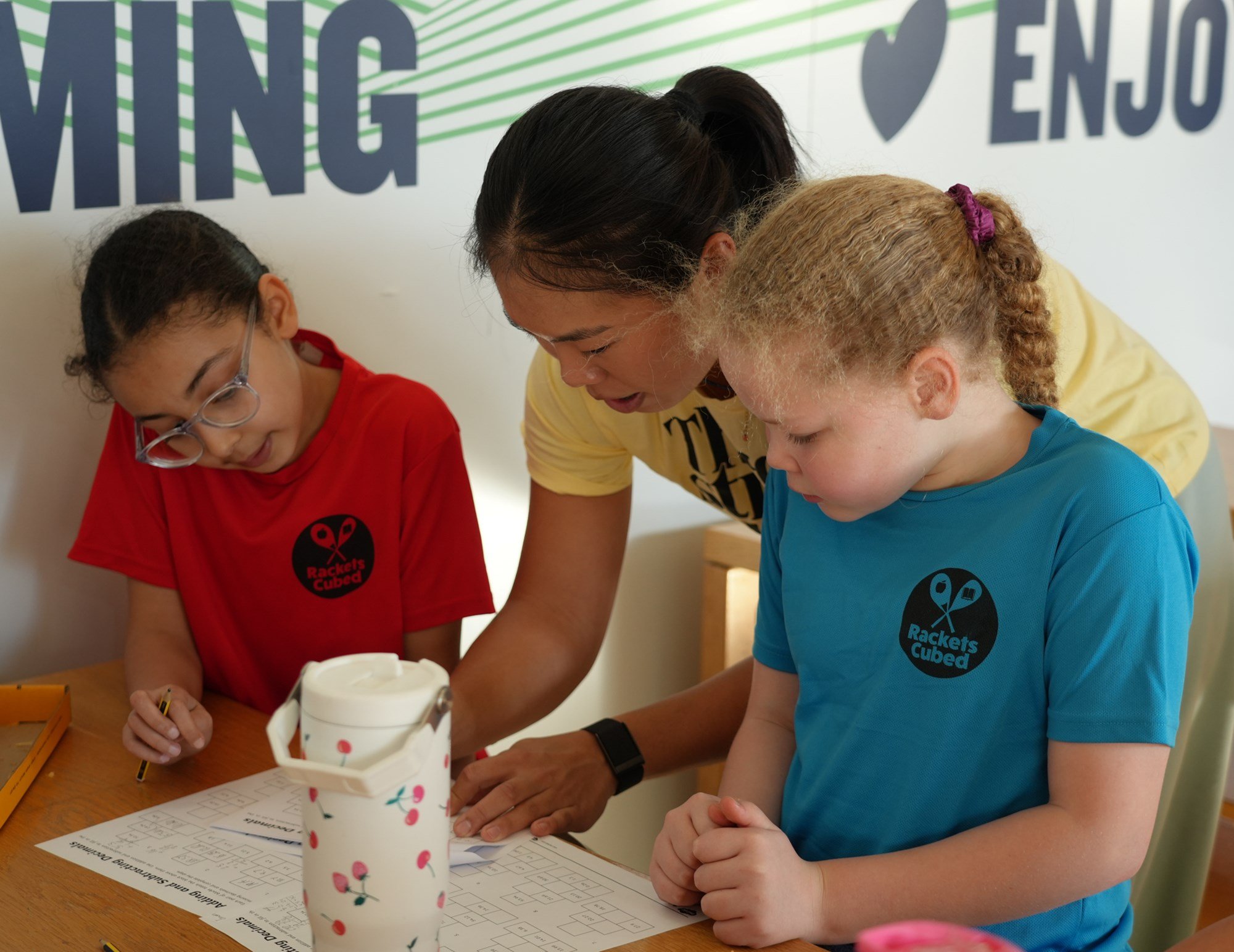 An older girl in a yellow t-shirt helps two young girls, in a blue and red t-shirt,with their homework