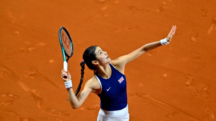 Emma Raducanu tossing a tennis ball in the air while preparing to serve on an orange clay court 