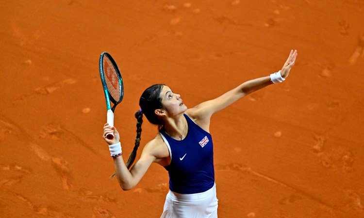 Emma Raducanu tossing a tennis ball in the air while preparing to serve on an orange clay court 