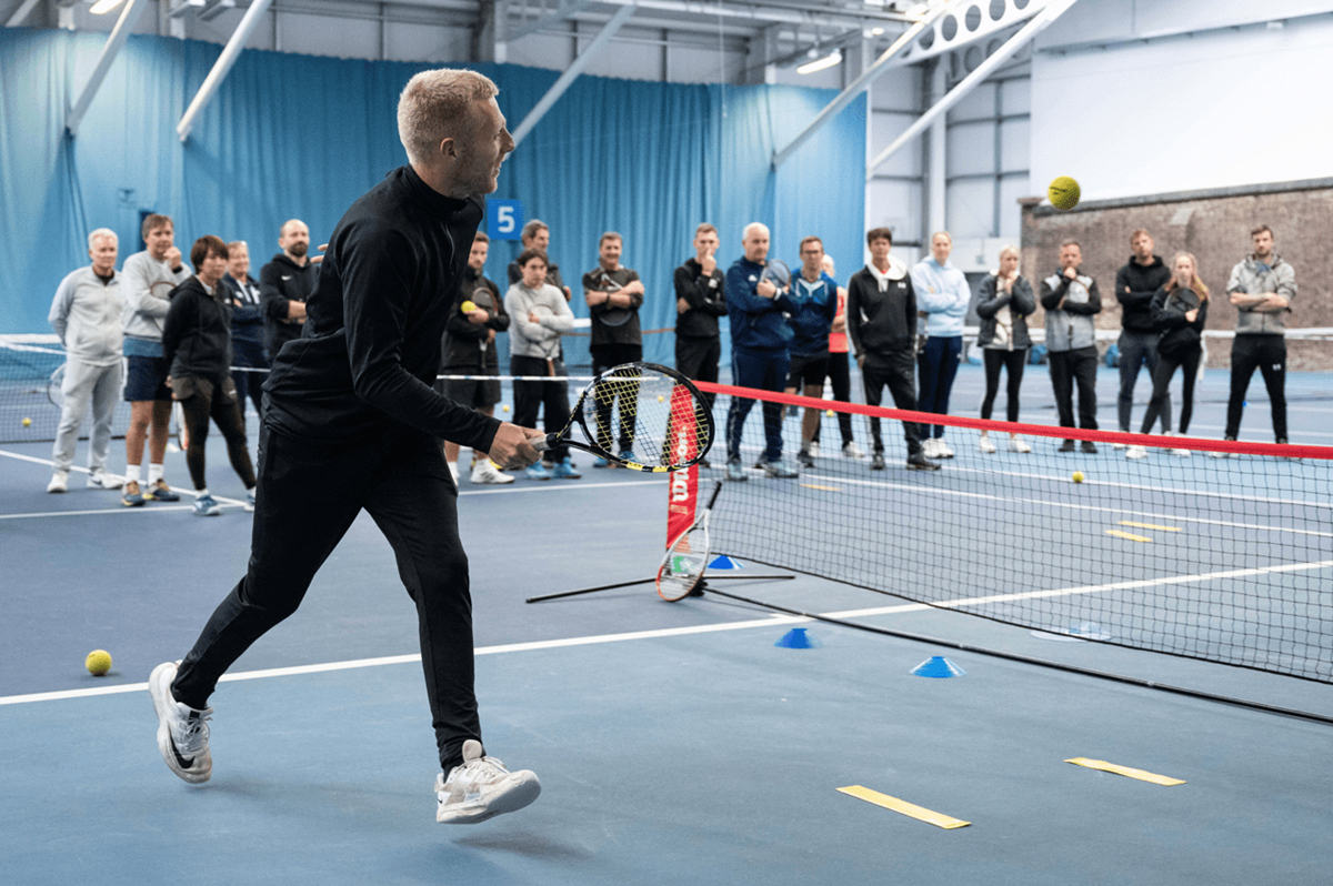 Prospective tennis coaches standing on a tennis court watching a tutor during an LTA on-court workshop