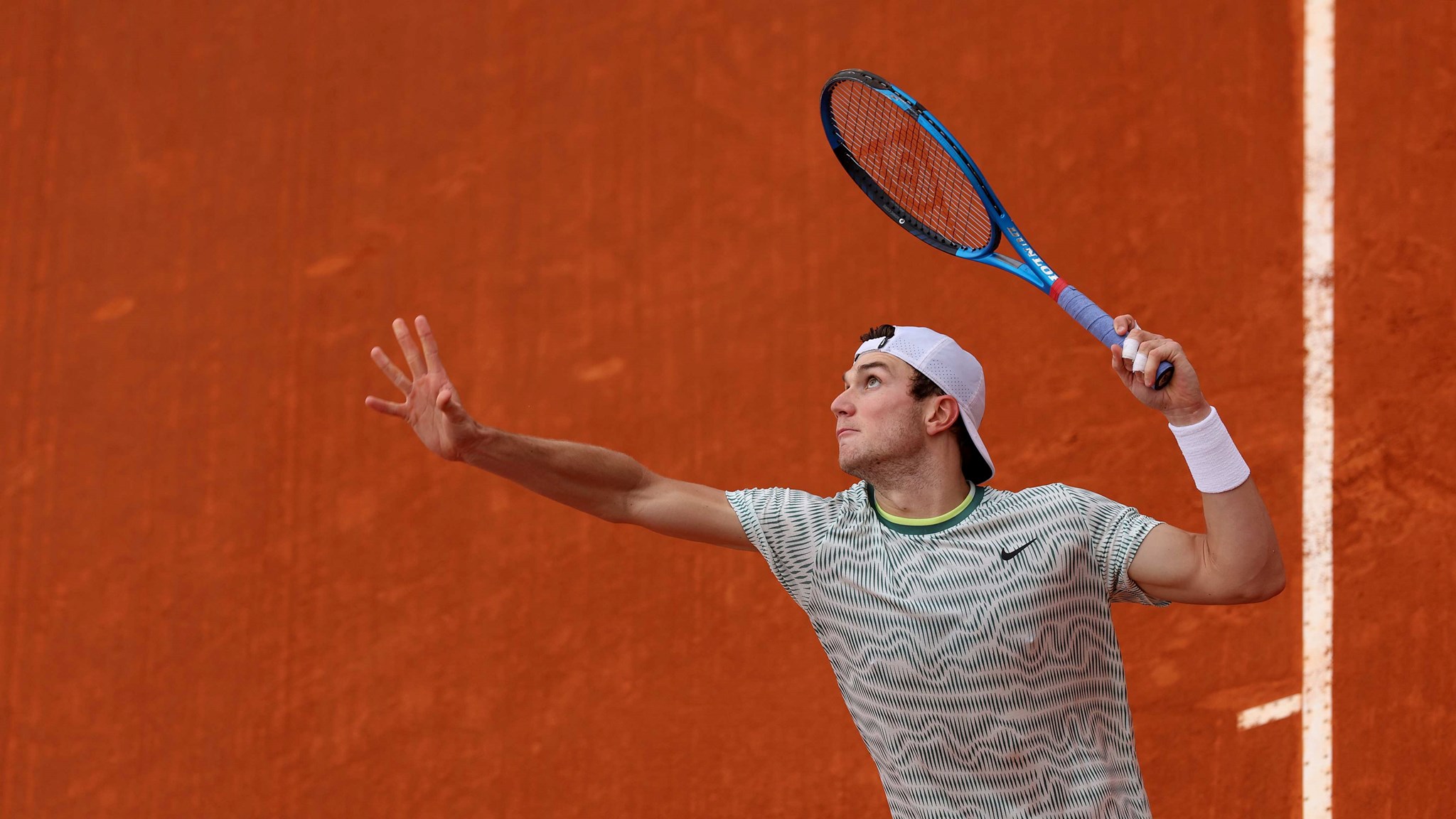 An image of Jack Draper mid-service motion with a clay court in the background