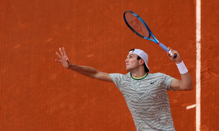 An image of Jack Draper mid-service motion with a clay court in the background