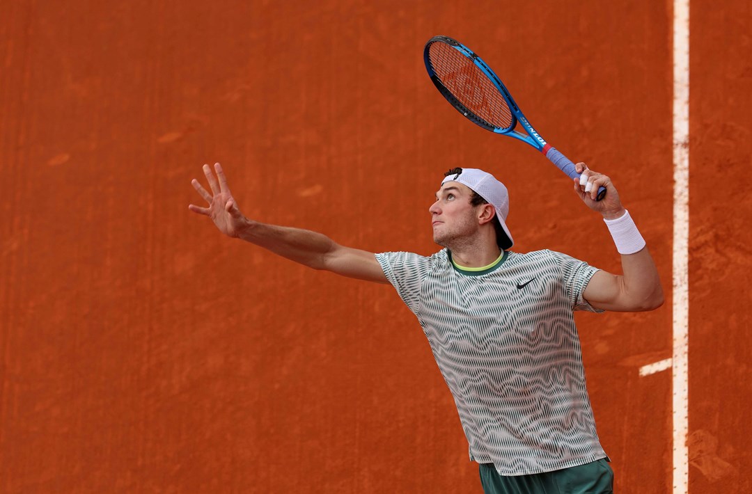 An image of Jack Draper mid-service motion with a clay court in the background