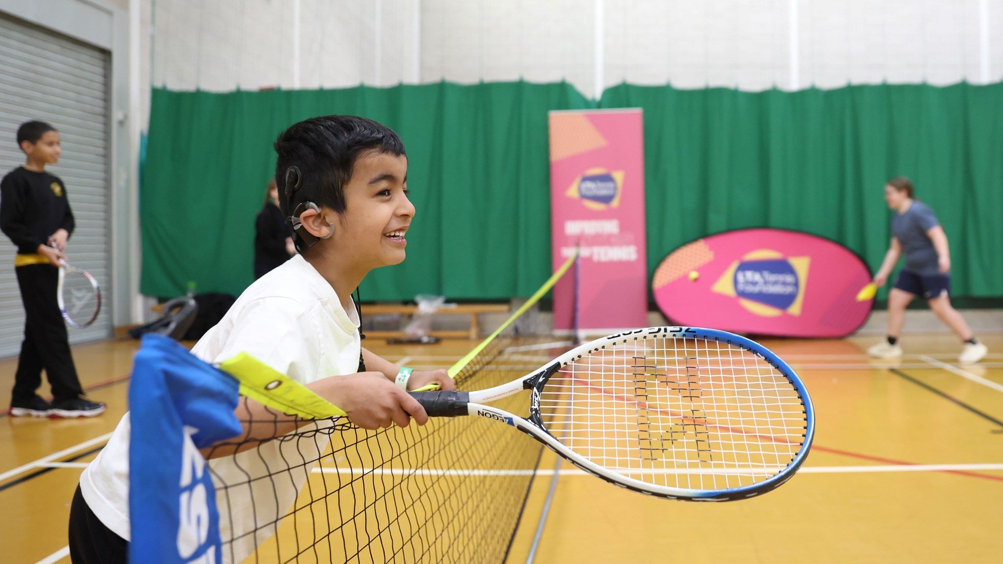 Young boy with a cochlear implant holding a tennis racket and leaning over the net