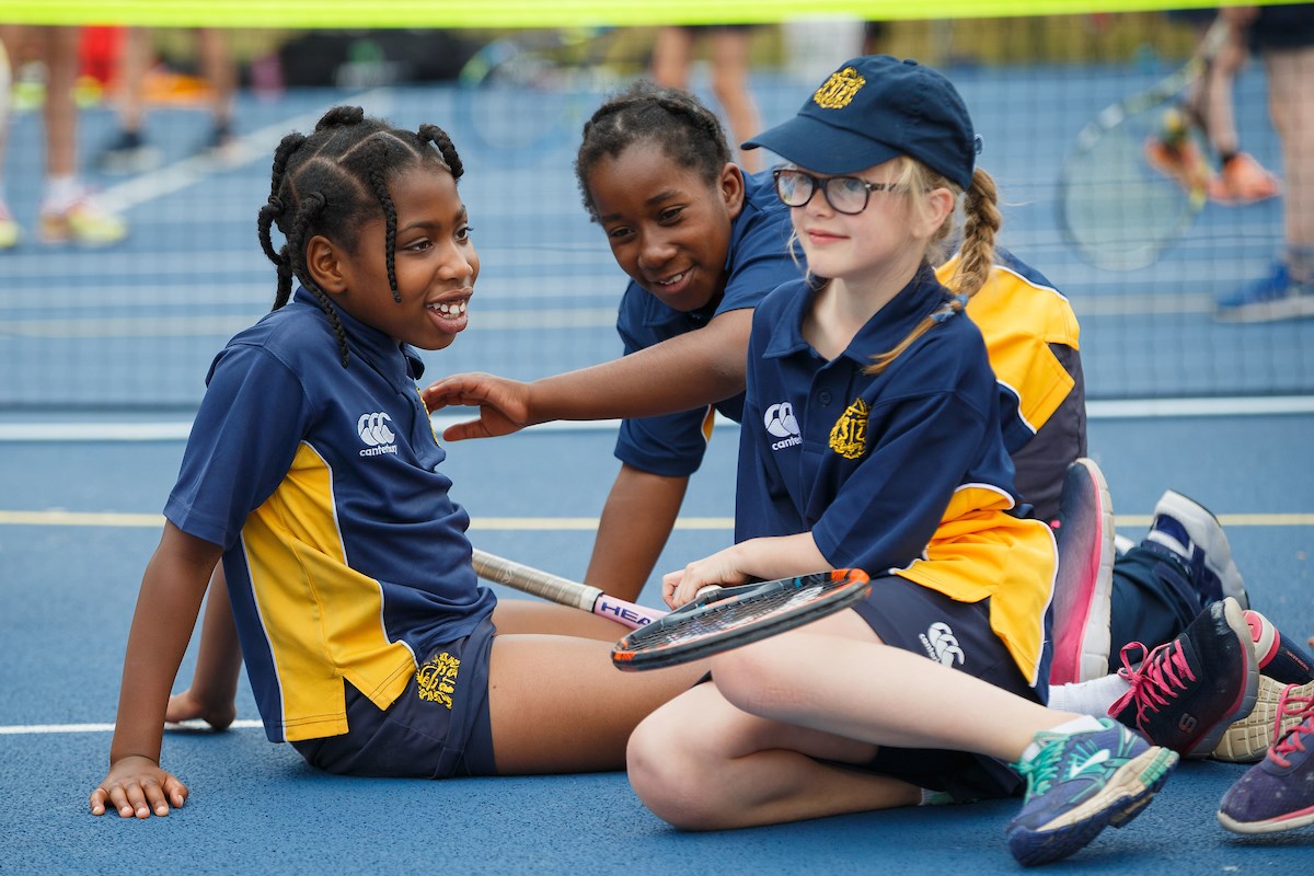 Three school children sat on a tennis court holding tennis rackets