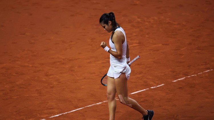 Emma Raducanu clenching her fist while stood on a clay court in a white tennis dress 
