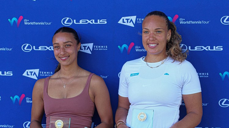 Two women smile whilst holding glass trophies. The women on the left is wearinng a brown vest top, and the women on the right is wearing a white t-shirt