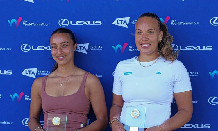Two women smile whilst holding glass trophies. The women on the left is wearinng a brown vest top, and the women on the right is wearing a white t-shirt