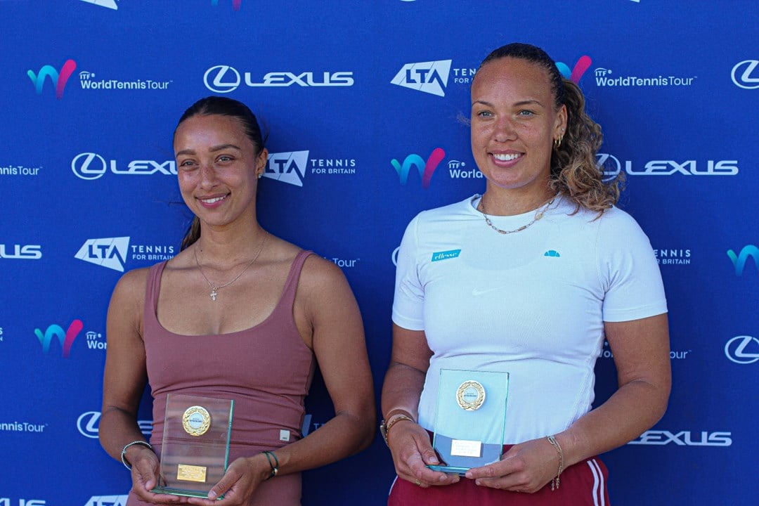 Two women smile whilst holding glass trophies. The women on the left is wearinng a brown vest top, and the women on the right is wearing a white t-shirt