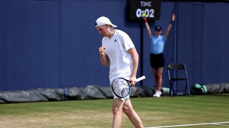A man wearing white sports gear clenches his fist in celebration, while holding a tennis racket in his other hand. He is on a tennis court and is turning to walk towards the back of the court; in the background, a girls wearing a light blue polo top and a black skirt holds her arms aloft, with a tennis ball in her right hand