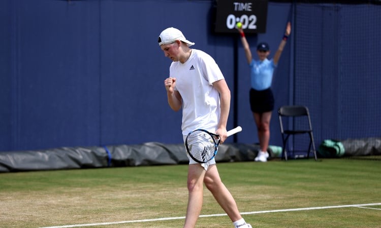 A man wearing white sports gear clenches his fist in celebration, while holding a tennis racket in his other hand. He is on a tennis court and is turning to walk towards the back of the court; in the background, a girls wearing a light blue polo top and a black skirt holds her arms aloft, with a tennis ball in her right hand