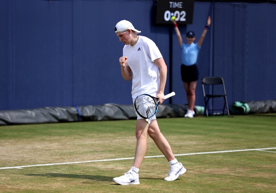 A man wearing white sports gear clenches his fist in celebration, while holding a tennis racket in his other hand. He is on a tennis court and is turning to walk towards the back of the court; in the background, a girls wearing a light blue polo top and a black skirt holds her arms aloft, with a tennis ball in her right hand