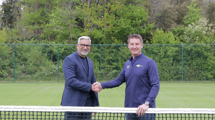 Two men standing behind a tennis net shaking hands and smiling. They are stood on grass, with green foliage in the background behind them