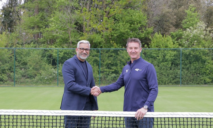 Two men standing behind a tennis net shaking hands and smiling. They are stood on grass, with green foliage in the background behind them