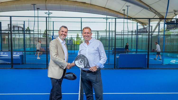Two men stand on a blue padel court, shaking each other's hand and each holding a padel bat in the other. People on another padel court are visible in the background.
