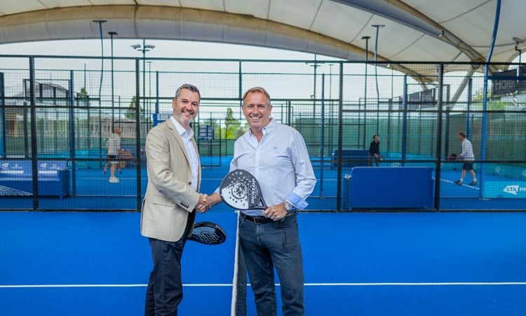 Two men stand on a blue padel court, shaking each other's hand and each holding a padel bat in the other. People on another padel court are visible in the background.