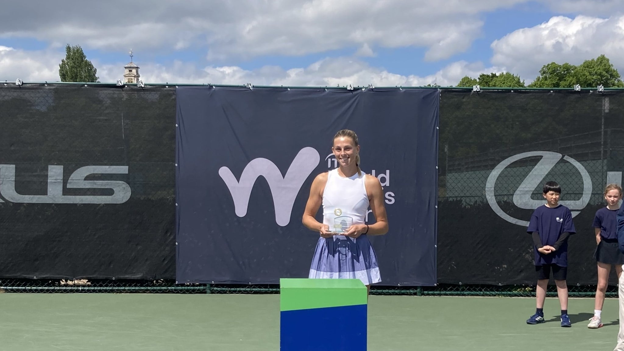 A woman wearing a white vest top and a lilac skirt holds a glass trophy, standing behind a blue pedestal with the words, 'LTA, Tennis for Britain' on it