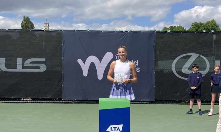 A woman wearing a white vest top and a lilac skirt holds a glass trophy, standing behind a blue pedestal with the words, 'LTA, Tennis for Britain' on it