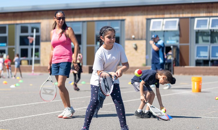 an image of a young girl holding a tennis racket and hitting a ball on court with other children and an older woman stood around her