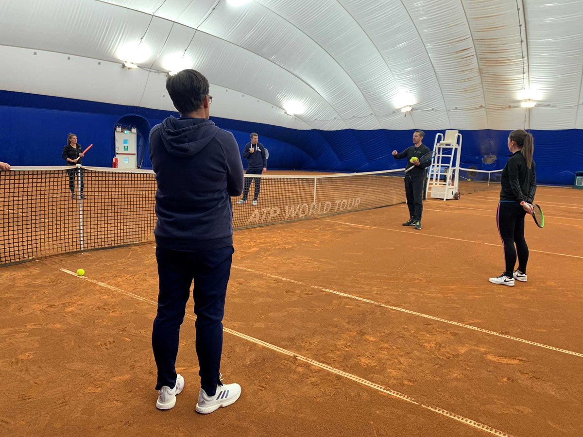 Tennis coach explaining a drill to prospective coaches on a clay court