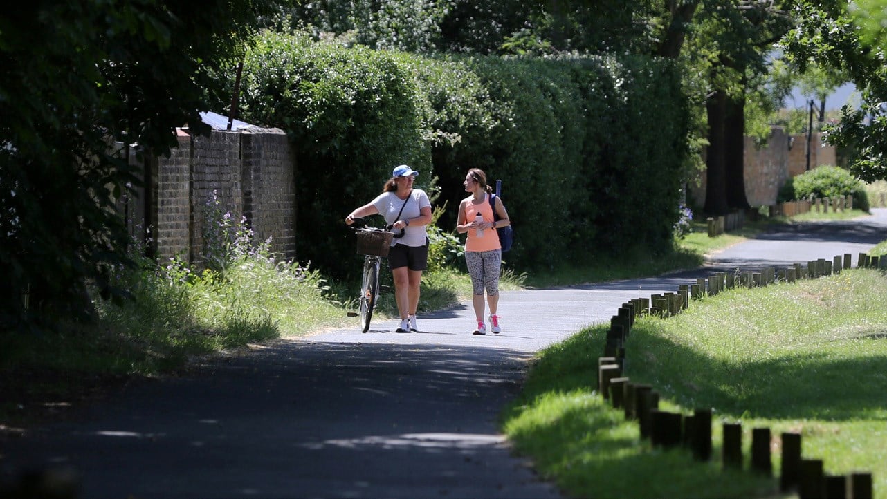 Two women walking on a tarmac path through a park, dressed in sports gear, with the lady on the left guiding a bicycle