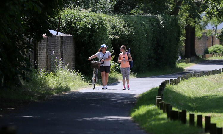 Two women walking on a tarmac path through a park, dressed in sports gear, with the lady on the left guiding a bicycle