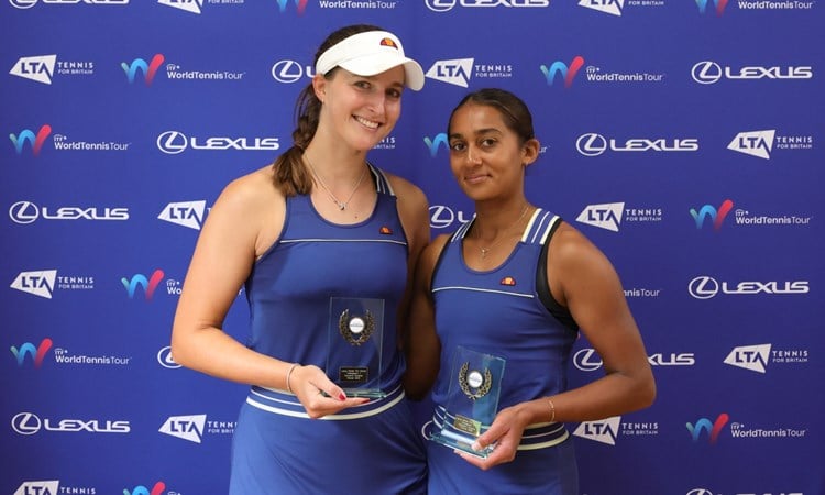 Two women wearing blue tennis dresses and holding glass trophies, stood in front of a blue background with sponsor logos from the LTA, ITF World Tennis Tour, and Lexus on display